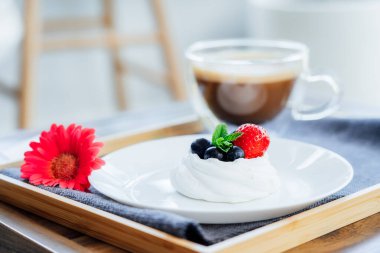 Pavlova tatlı mini kek, bir fincan latte kahve ve masa üzerinde gri peçete olan ahşap tepside taze gerbera çiçeği. Kahve zamanı. Sevdiğin için kahvaltı. Biraz ara ver ve rahatla. Seçici odak.