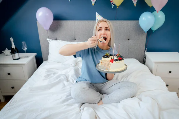 Smiling woman in pajama and party cap eating birthday cake by spoon, sitting on the bed in decorated bedroom. Morning surprise, fun during home party. Happy birthday concept. Selective focus.