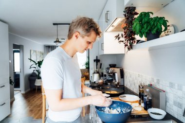 Genç bir adam evdeki modern bir hafif mutfakta peynirli krep pişiriyor. Adam tavada kızarmak için krep hazırlıyor. Ev işleri ve ev işleri. Mutfak hobisi. Seçici odak.