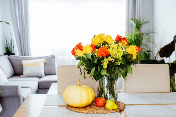 A vase of yellow and orange roses flowers, fresh pumpkin, apple and pear on a kitchen table counter with open space living room background. Autumn home decor for Thanksgiving and Halloween holiday