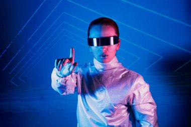 Neon portrait of woman, dressed in futuristic holographic clothes and glasses touch space by finger. Cyberpunk girl with virtual touching pad. Neon blue and pink light. Virtual reality glasses