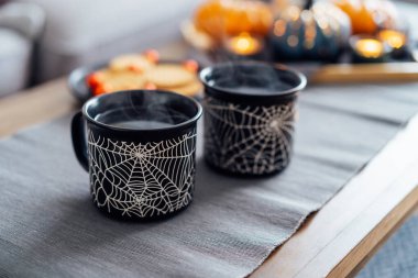 Cozy halloween plans at home. Hot tea drink in black mugs with spider net pattern, cookies and and sweets on the plate, pumpkin decor on the coffee table. Festive autumn, fall mood home decor