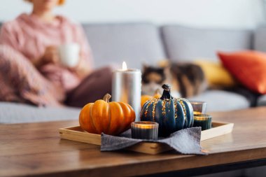 Autumn, fall cozy mood composition for hygge home decor. Small pumpkins, burning candles on tray with gray napkin on the coffee table with resting woman with cat in the living room. Selective focus.