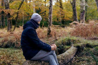 Young man in warm clothes sitting on a fallen tree near river, looking at the forest and enjoying the moment. Feeling harmony, reunion with nature in autumn. Relaxing, personal fulfillment.