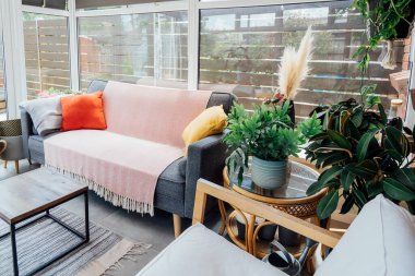 Parlak tekstil örtüsü, yastıklar ve bir sürü yeşil ev bitkisiyle dekore edilmiş gri koltuklu rahat, hafif konservatuar iç tasarımı. Hygge ev dekorasyonu. Biyofilik yaşam tarzı. Seçici odak.