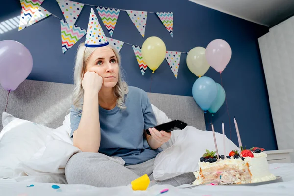 Sad woman in pajamas, party cap drinks champagne and watches TV, wrapped in a blanket while sitting on a bed in a decorated bedroom. Celebrates a birthday alone. Boring home party. Selective focus.