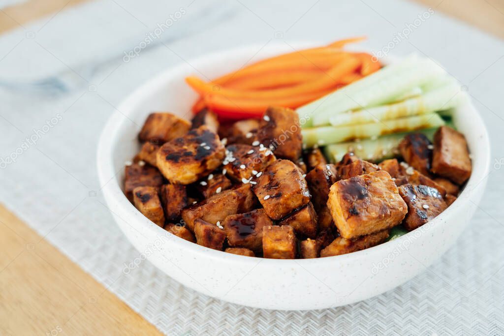 Bowl with roasted tempeh made of fermented soy beans, fresh vegetable sticks with sauce and sesame seeds on the table. Plant based protein. Healthy cooking and eating. Go vegan. selective focus.