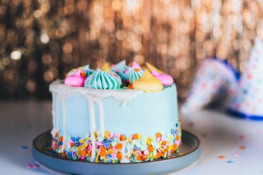Colorful birthday cake with sprinkles and festive caps on the sparkling gold tinsel background. Festive birthday celebration, party. Selective focus, copy space.