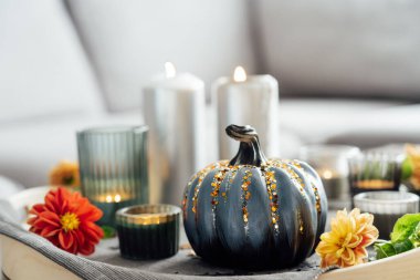 Autumn cozy mood composition for hygge home decor. Gray pumpkin with sequins, candles and fresh dahlia flowers on the tray with gray napkin on the coffee table in the living room. Selective focus.