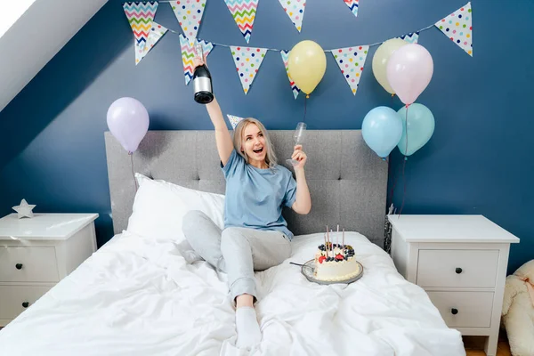 Happy emotional woman in pajama and party cap sitting on the bed with festive cake and champagne bottle in decorated bedroom. Time for yourself Celebration at home. Happy birthday concept