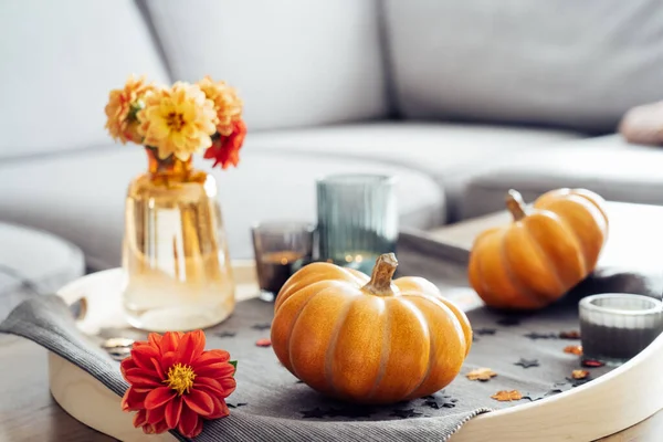 Autumn, fall cozy mood composition for hygge home decor. Small pumpkins, burning candles and fresh dahlia flowers on the tray with gray napkin on the coffee table in the living room. Selective focus.