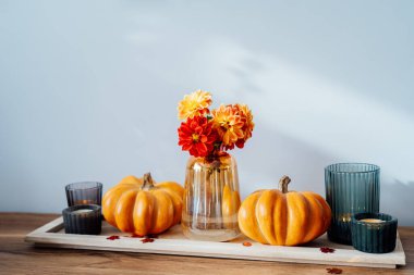 Autumn, fall cozy composition. Orange pumpkins, candles, dahlia flowers in vase on the wooden tray on the table with white wall background. Scandinavian minimalist hygge home decor. Selective focus