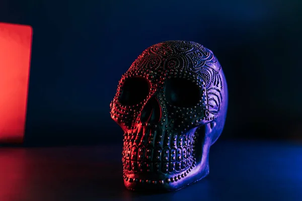 Black human skull decorated with golden points pattern in neon purple and red light on dark blue background. Halloween decor concept. Selective focus, copy space.