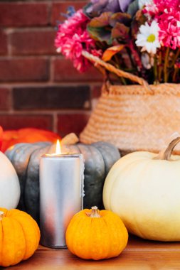 Festive composition for autumn holidays. Burning candle, various pumpkins and flowers bouquet in the wicker basket on brik wall background. Thanksgiving or Halloween. Fall Harvesting. Selective focus.