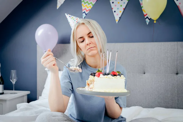 Sad, upset woman in pajama and party cap thinking eat or not to eat birthday cake sitting on the bed in decorated bedroom. Celebrates birthday alone concept. Selective focus.