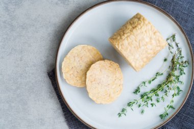 Top view raw Tempeh or Tempeh, made of fermented soybean seeds. Tempeh Slices on plate with herbs. Soya protein. Plant based meat concept. Healthy Cooking and eating. Go vegan. Flat Lay, copy space.