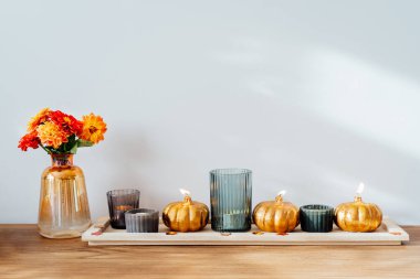 Autumn, fall cozy composition. Fresh dahlia flowers in vase and pumpkin shaped golden candles on the wooden tray with white wall background. Scandinavian minimalist hygge home decor. Selective focus.