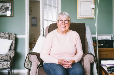 Portrait of smiling senior, mature retired grandmother in eyeglasses, elderly woman smiling, sitting on the armchair at home enjoying retirement. Selective focus