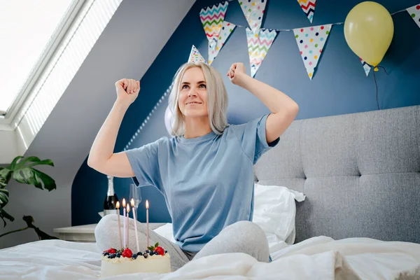 Smiling emotional woman in birthday cap with festive cake with candles dancing on the bed in decorated bedroom. Morning surprise. Happy birthday concept. Selective focus.