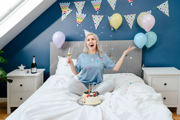 Happy emotional woman in pajama and party cap with champagne glass throwing confetti up while sitting on the bed in a decorated bedroom.Time for yourself Celebration at home. Happy birthday concept.