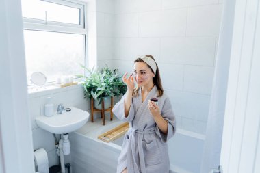 Young woman in bathrobe looking in the mirror and applying facial natural cosmetic clay mask on her face in bathroom. Cosmetic procedures for skin care at home. Beauty self-care. Selective focus.