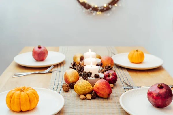 Fall table setting for celebration Thanksgiving day family party. Autumn composition with candles, fruits, nuts and cones. Plates with pumpkins and pomegranate. Natural autumn decor. Selective focus.