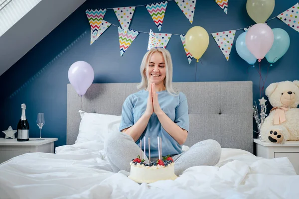 Happy emotional woman in pajama and party cap after blowing out the candles on birthday cake, sitting on the bed in decorated bedroom. Make a wish process. Morning surprise. Happy birthday concept