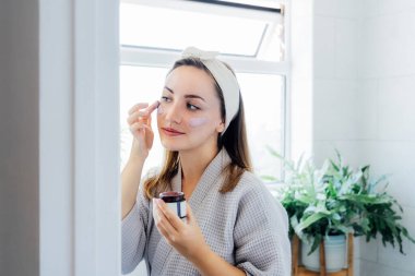 Young woman in bathrobe looking in the mirror and applying facial natural cosmetic clay mask on her face in bathroom. Cosmetic procedures for skin care at home. Beauty self-care. Selective focus
