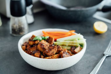 Bowl with roasted tempeh made of fermented soy beans, fresh vegetable sticks with sauce and sesame seeds on the table. Plant based protein. Healthy cooking and eating. Go vegan. selective focus.