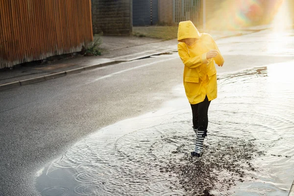 雨の後 女性は通りで楽しんでいます 雨のゴムブーツと黄色のレインコートを着て笑顔の女性は水スプラッシュと水たまりに歩いて日没の光の中でドロップします 選択的焦点 — ストック写真