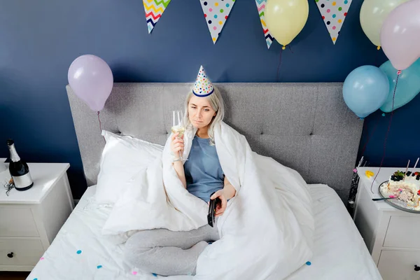 Sad woman in pajamas, party cap drinks champagne and watches TV, wrapped in a blanket while sitting on a bed in a decorated bedroom. Celebrates a birthday alone. Boring home party. Selective focus.