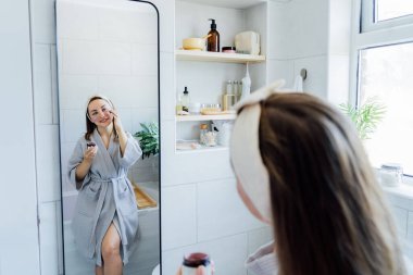 Young woman in bathrobe looking in the mirror and applying facial natural cosmetic clay mask on her face in bathroom. Cosmetic procedures for skin care at home. Beauty self-care. Selective focus