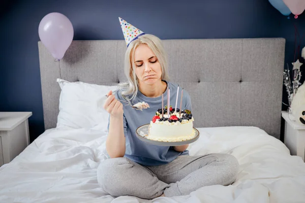 Sad, upset woman in pajama and party cap thinking eat or not to eat birthday cake sitting on the bed in decorated bedroom. Celebrates birthday alone concept. Selective focus.