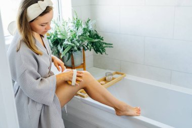 Bornozlu genç bir kadın açık renkli banyoda sabah duşundan önce kuru fırçayla masaj yapıyor. Anti-selülit tedavisi, lenfatik drenaj masajı. Kadın güzeli. Seçici odak