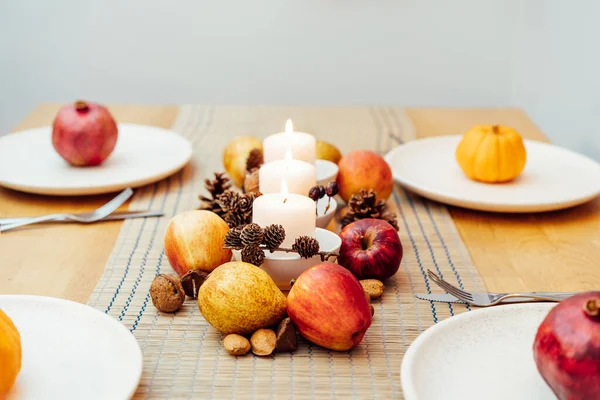 Fall table setting for celebration Thanksgiving day family party. Autumn composition with candles, fruits, nuts and cones. Plates with pumpkins and pomegranate. Natural autumn decor. Selective focus.