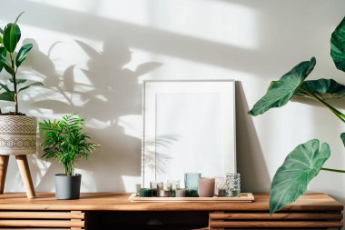 Modern minimalist İskandinav tarzı iç tasarımı beyaz poster modeli, mumlar ve tropikal yeşil ev bitkileri güneş ışığı altında ve gri bir duvarda gölgeler. Seçici odak.