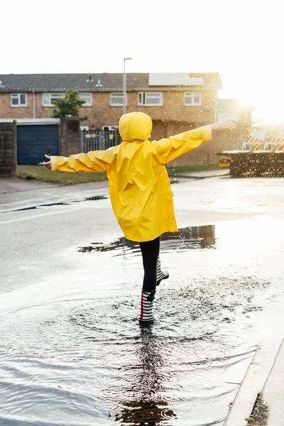 雨の後 女性は通りで楽しんでいます バックビューの女性は 雨のゴムブーツと黄色のレインコートの水スプラッシュと水たまりに歩いて着て 日没の光の中でドロップします 選択的フォーカス — ストック写真