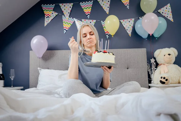 Sad, upset woman in pajama and party cap thinking eat or not to eat birthday cake sitting on the bed in decorated bedroom. Celebrates birthday alone concept. Selective focus.