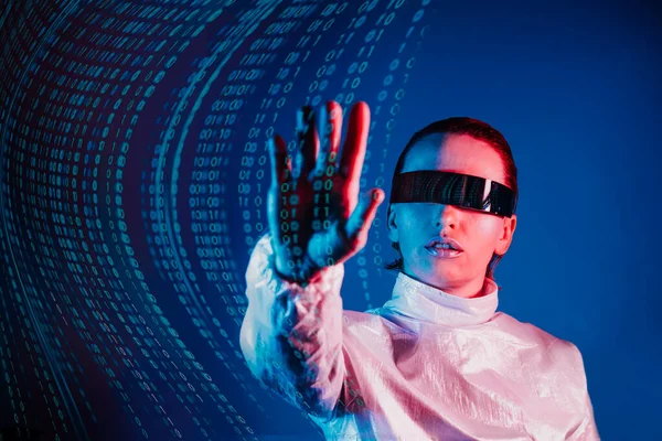 Woman dressed in holographic clothes and futuristic eyeglasses with reflection of matrix code is touching by hand matrix digital numbers artificial intelligence data before her in blue light.