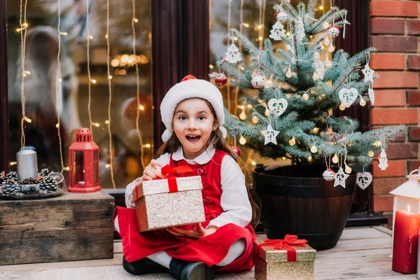 Excited and surprised cute girl kid in red dress and Santa hat holding gift box present sitting on the house porch with decorated Christmas tree. Christmas holiday season. Selective focus