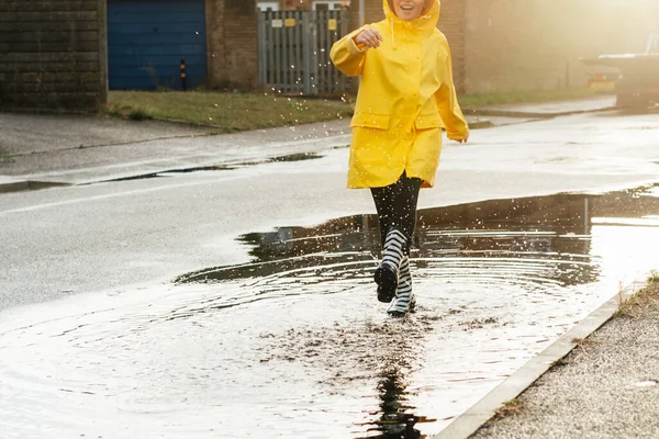 雨の後 女性は通りで楽しんでいます 雨のゴムブーツと黄色のレインコートを身に着けているトリミングされた女性は 水のスプラッシュと日没の光のドロップで水たまりに歩いてください 選択的焦点 — ストック写真