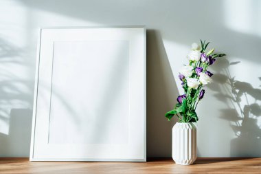Modern minimalist İskandinav tarzı iç tasarımı beyaz poster modeli ve vazodaki östoma çiçekleri güneş ışığı altındaki ahşap bir konsolda ve ev bitkilerinin gölgeleri beyaz gri bir duvarda. Seçici odak.