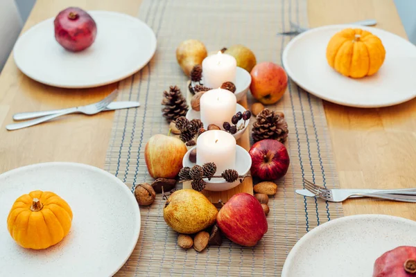 Fall table setting for celebration Thanksgiving day family party. Autumn composition with candles, fruits, nuts and cones. Plates with pumpkins and pomegranate. Natural autumn decor. Selective focus.