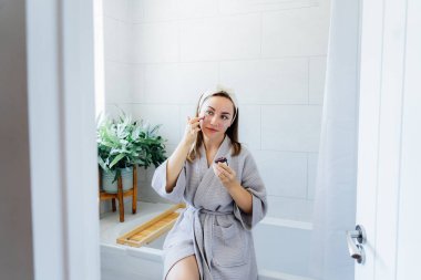 Young woman in bathrobe looking in the mirror and applying facial natural cosmetic clay mask on her face in bathroom. Cosmetic procedures for skin care at home. Beauty self-care. Selective focus