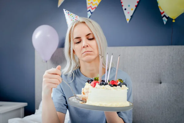 Sad, upset woman in pajama and party cap thinking eat or not to eat birthday cake sitting on the bed in decorated bedroom. Celebrates birthday alone concept. Selective focus.