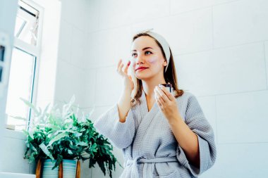 Young woman in bathrobe looking in the mirror and applying facial natural cosmetic clay mask on her face in bathroom. Cosmetic procedures for skin care at home. Beauty self-care. Selective focus