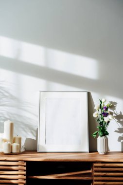Modern minimalist İskandinav tarzı iç tasarımı beyaz poster modeli, mum ve çiçekler, güneş ışığı altındaki ahşap bir konsolda ve ev bitkilerinin gölgeleri beyaz gri bir duvarda. Seçici odak.
