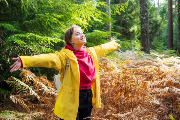 明るい黄色のレインコートで腕を上げて笑顔の女性は 自然の中で秋の森の中での生活と自由を楽しんでいます 調和を感じ 秋には自然との再会 リラックスした個人的な充実感 — ストック写真