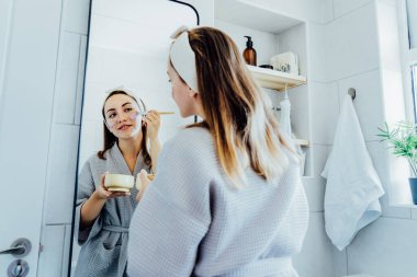 Young woman in bathrobe looking in the mirror and applying facial natural cosmetic clay mask on her face in bathroom. Cosmetic procedures for skin care at home. Beauty self-care. Selective focus.