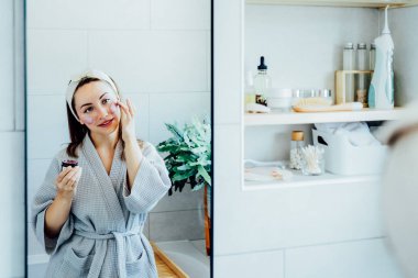 Young woman in bathrobe looking in the mirror and applying facial natural cosmetic clay mask on her face in bathroom. Cosmetic procedures for skin care at home. Beauty self-care. Selective focus.
